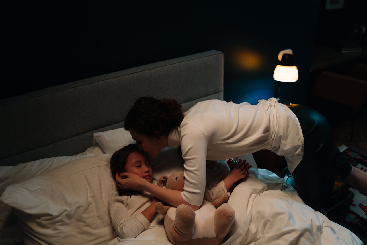 A mother tenderly kisses her sleeping child goodnight in a warmly lit bedroom, creating a peaceful scene.